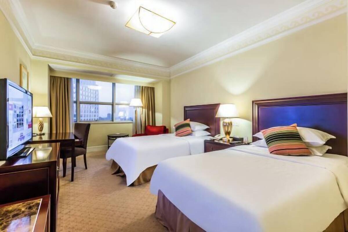 Ein Hotelzimmer mit zwei Betten, einem Schreibtisch mit Fernseher und einem Fenster mit Vorhängen.