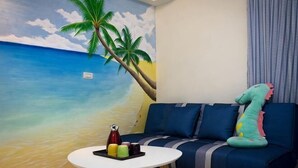 A room with a beach mural, a blue sofa, a small round table with a bottle and glasses, and a plush toy on the sofa. - Booksnwoods (Taitung)