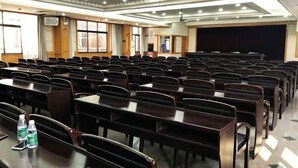 Une salle de conférence avec des rangées de tables et de chaises, un écran de projection et une estrade.