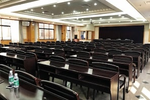 A conference room with rows of tables and chairs, a projector screen, and a stage area. - Ronglian Hotel (Foshan)