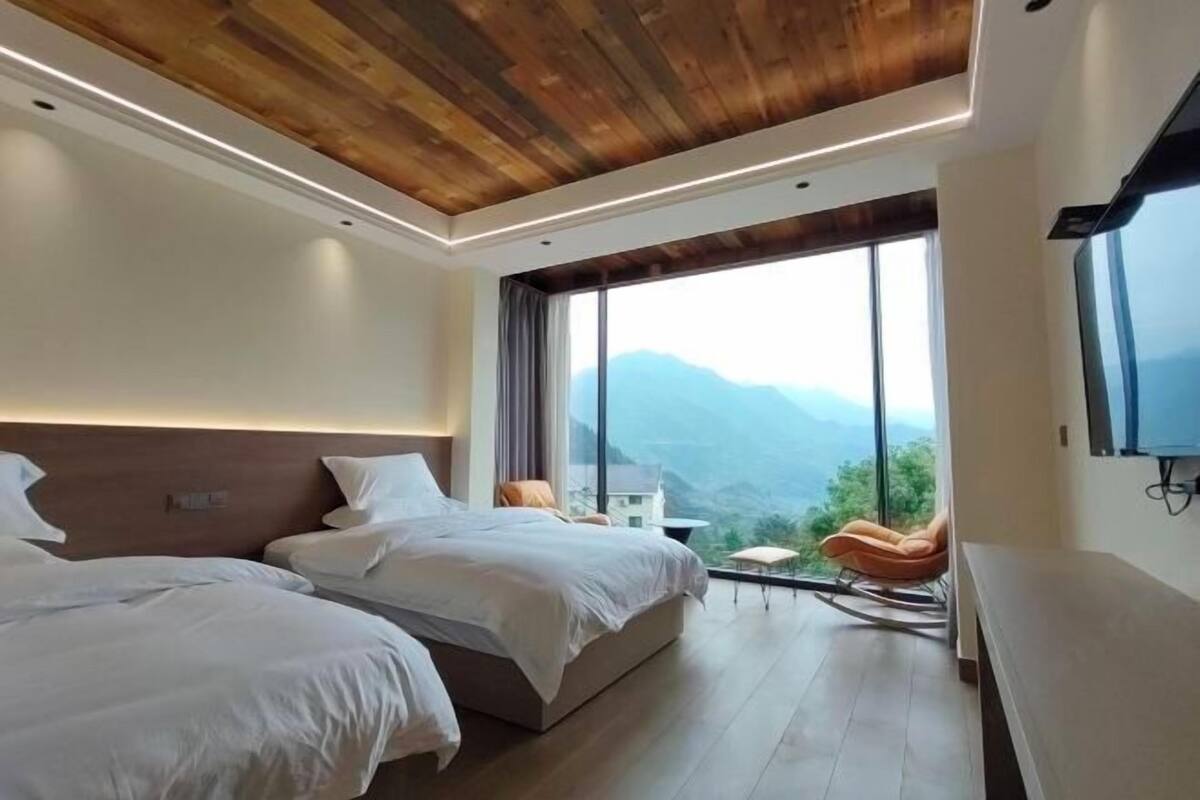 Una habitación de hotel moderna con dos camas, techo de madera, ventanas grandes y vistas a montañas.