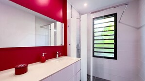 Ein modernes Badezimmer mit roter Akzentwand, einem großen Spiegel, einem Waschbecken mit rotem Seifenspender und einem Fenster mit horizontalen Jalousien.