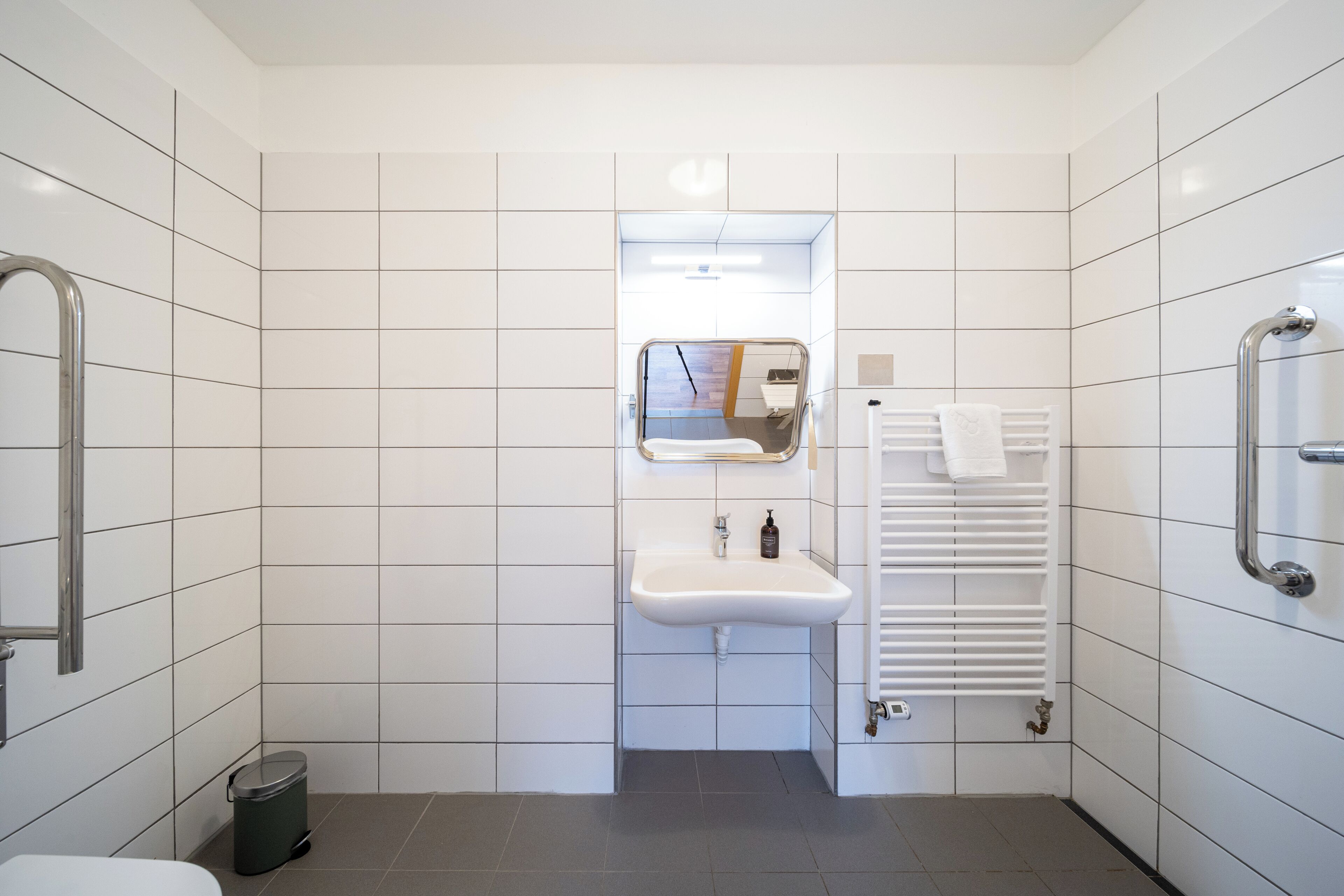 Un bagno moderno rivestito di piastrelle bianche, dotato di lavabo, specchio, porta asciugamani e zona doccia.