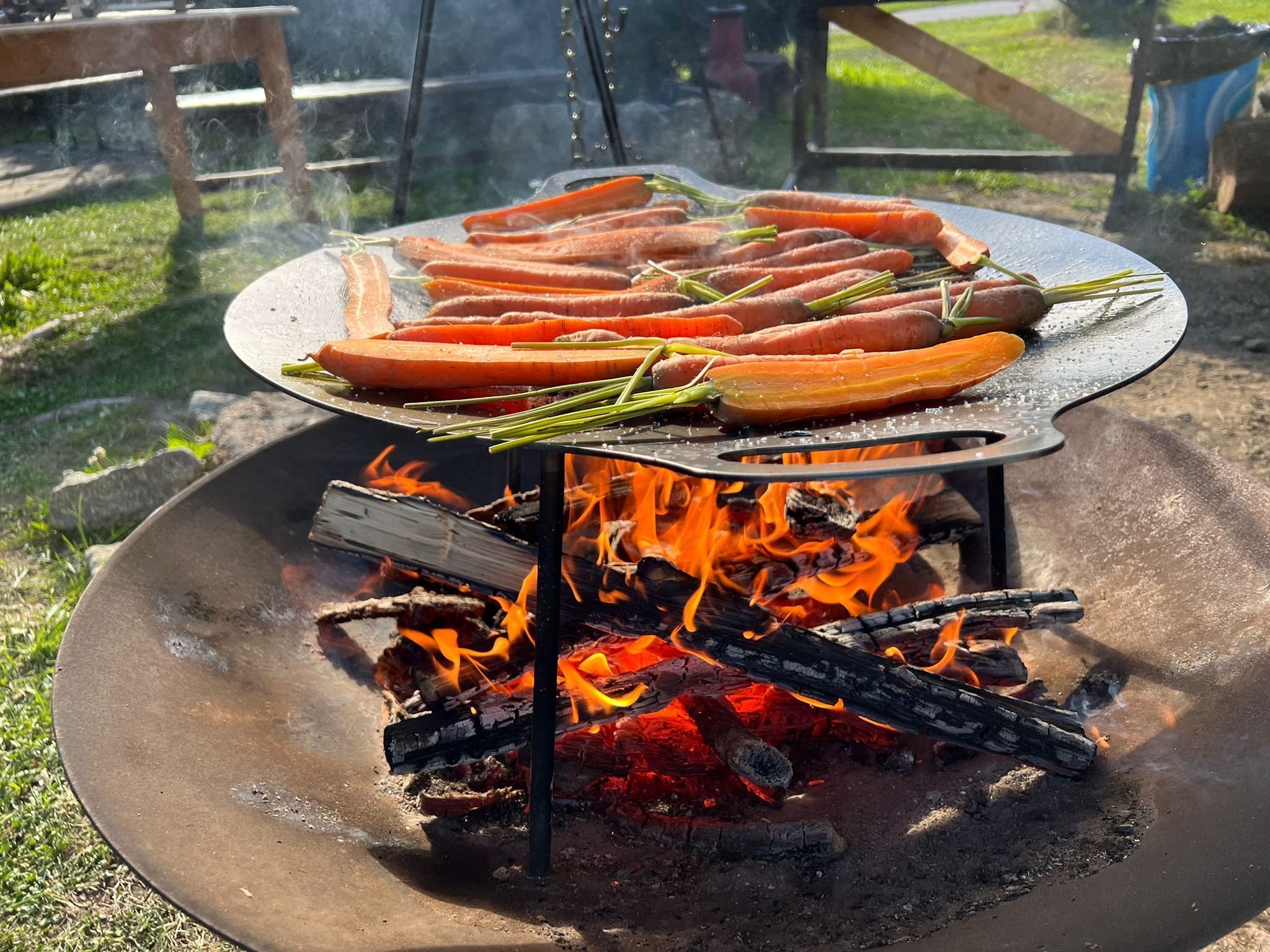 Una parrilla al aire libre circular con salchichas y verduras cocinándose sobre fuego abierto.