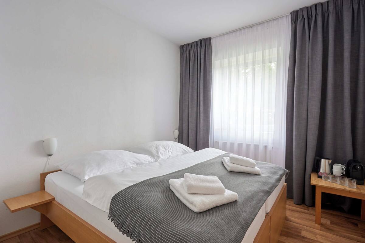 A neatly made bed with white linens and a gray throw, a bedside table with a lamp, and a window with gray curtains.
