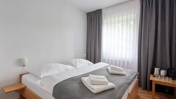Una cama bien hecha con sábanas blancas y una manta gris, una mesita de noche con lámpara y una ventana con cortinas grises.