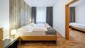 A modern hotel room with a wooden floor, a large bed with white linens, a bedside table with a lamp, and a view of the city through the window. - Resort Pasečná (Prední Výton)