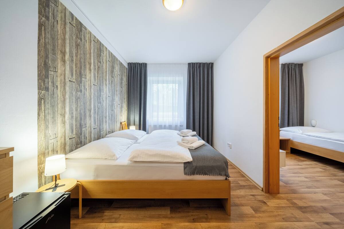 A modern hotel room with a wooden floor, a large bed with white linens, a bedside table with a lamp, and a view of the city through the window.