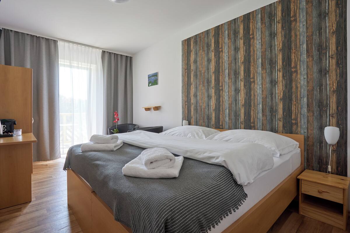 A neatly made bed with white linens, a grey blanket, and a folded towel. There is a wooden nightstand with a lamp beside the bed. The room has a wooden floor and a large window with grey curtains.