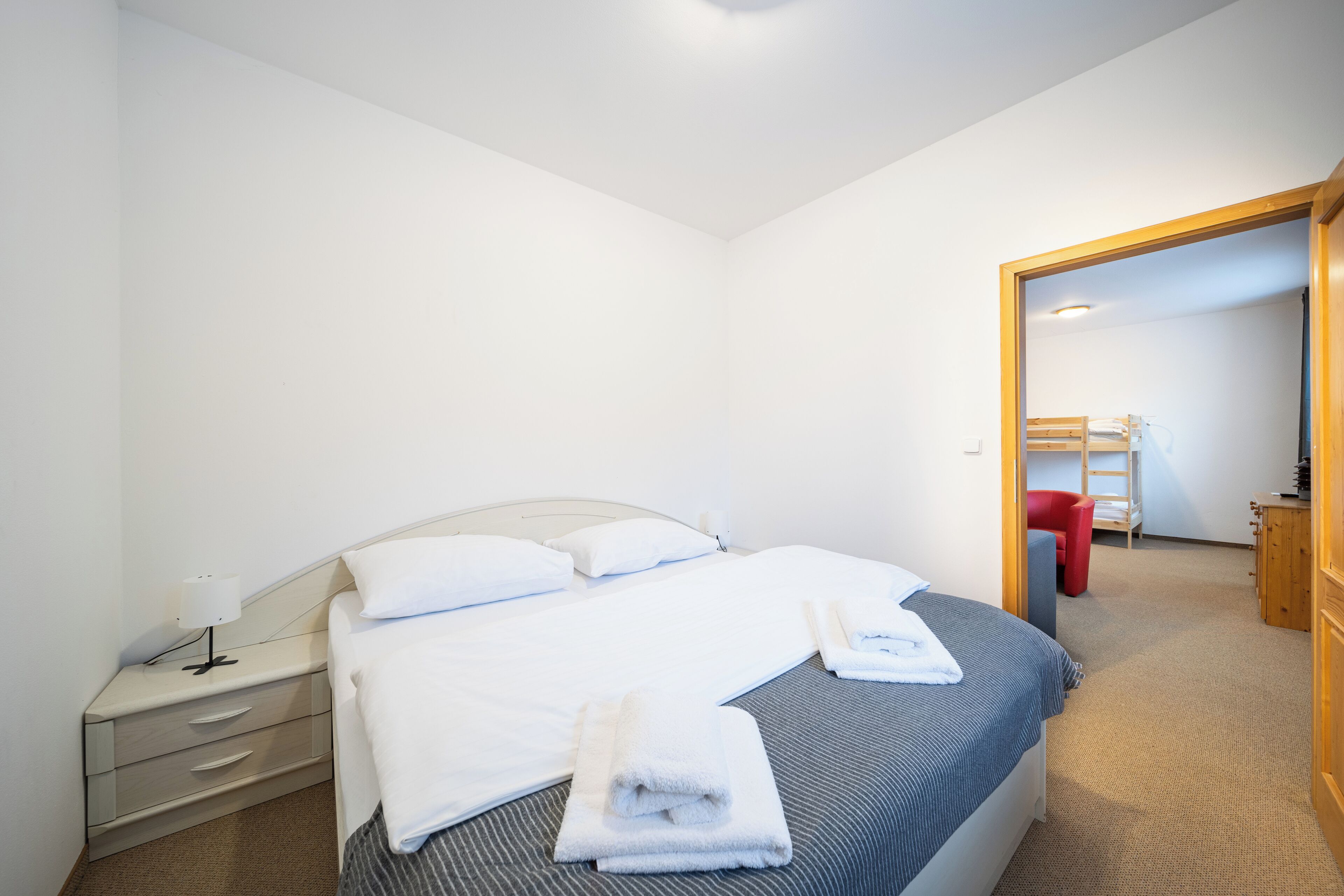 Una cama bien hecha con sábanas blancas y dos toallas dobladas, una mesita de noche con una lámpara, y una silla roja al fondo.