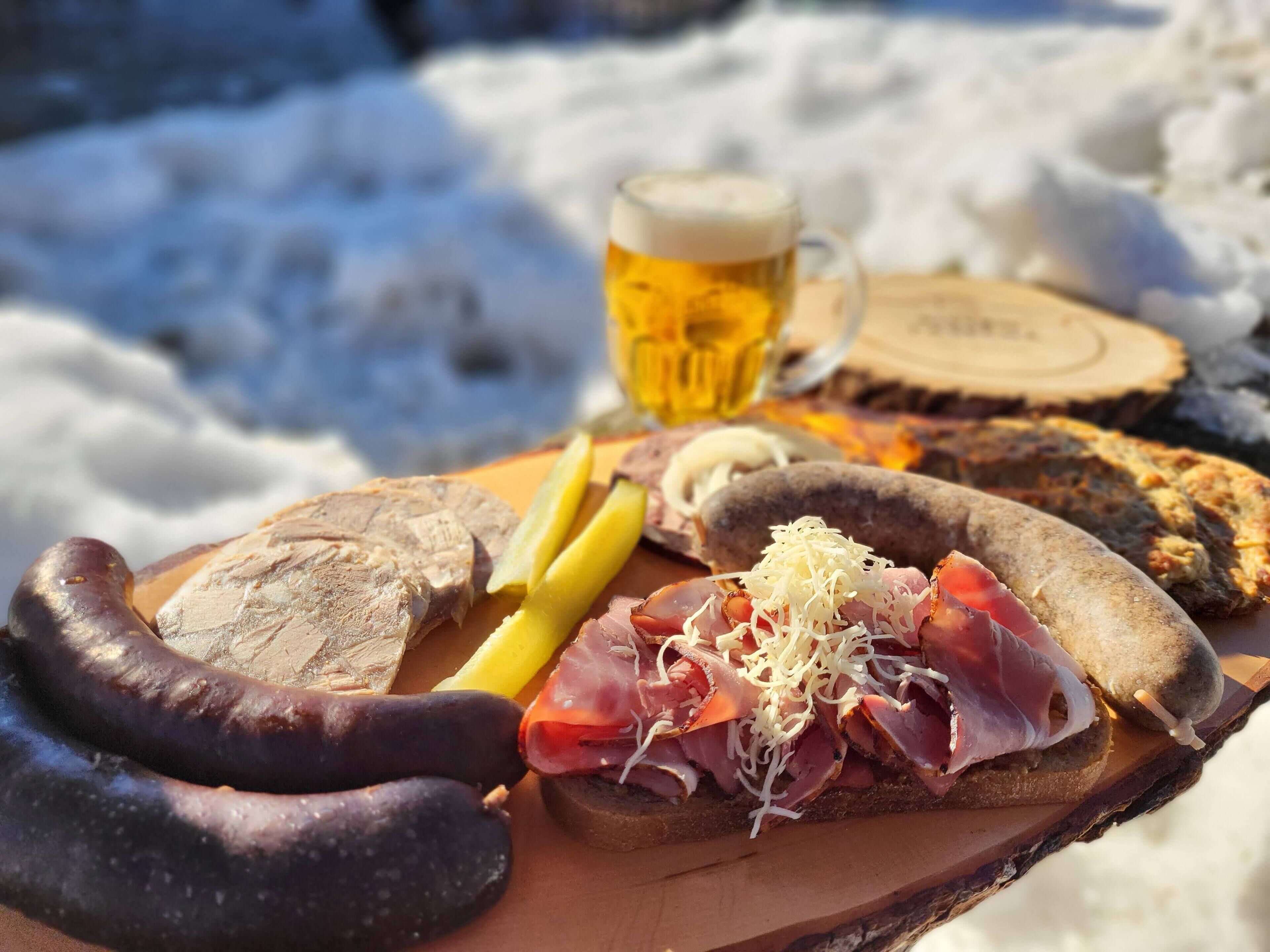 Une planche en bois garnie de différentes sortes de charcuterie, de cornichons et accompagnée d’une chope de bière, le tout présenté dans un décor extérieur enneigé.