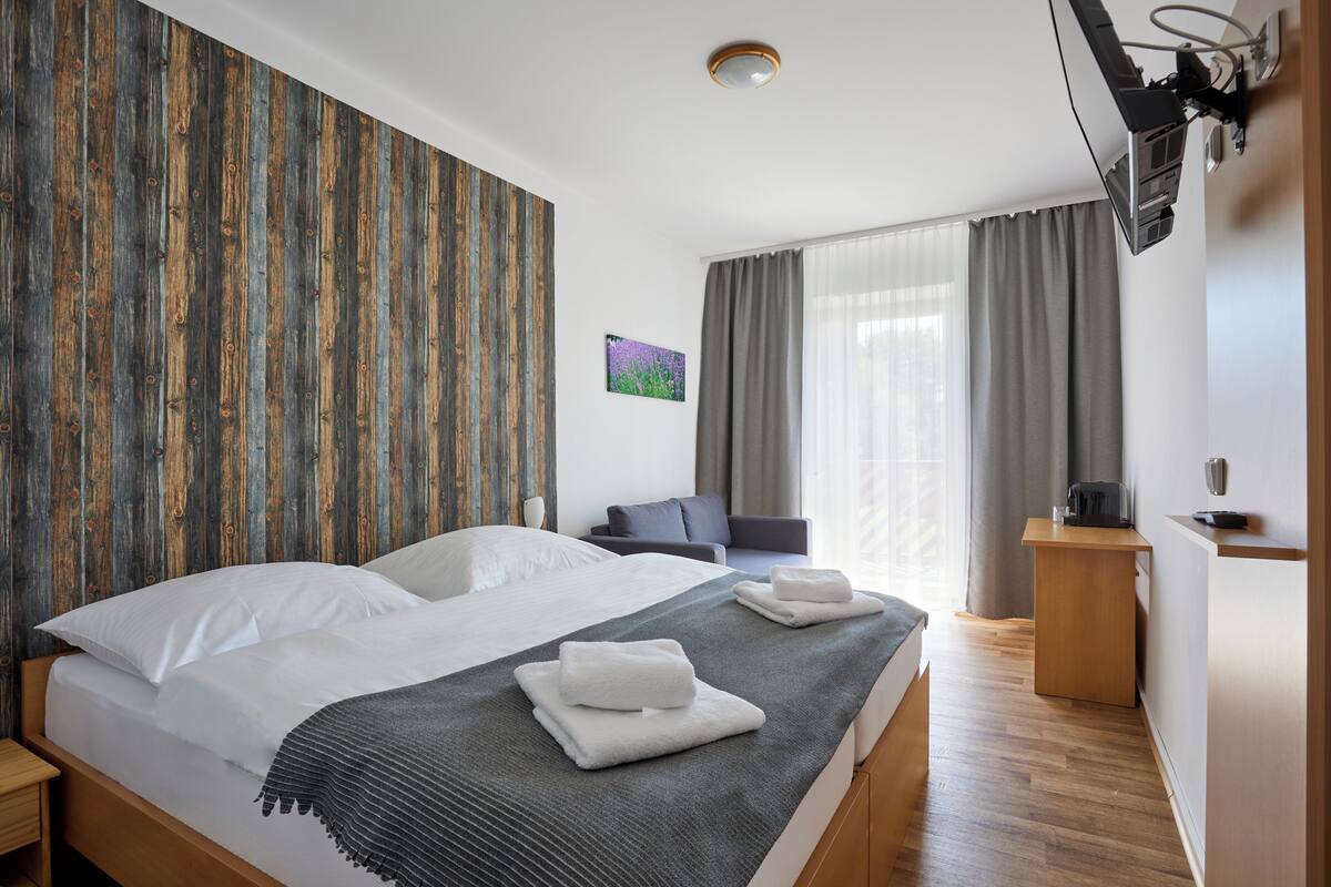 A hotel room with a wooden headboard, a bed with white linens, a grey throw blanket, a small bedside table, a desk with a lamp, and a window with curtains.