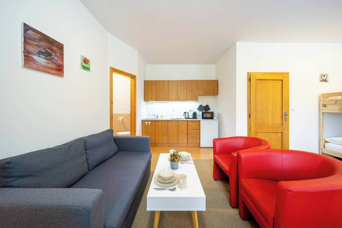 A modern living room with a grey sofa, a white coffee table with plates and a mug, and red armchairs. A kitchen with wooden cabinets and a microwave is visible in the background.
