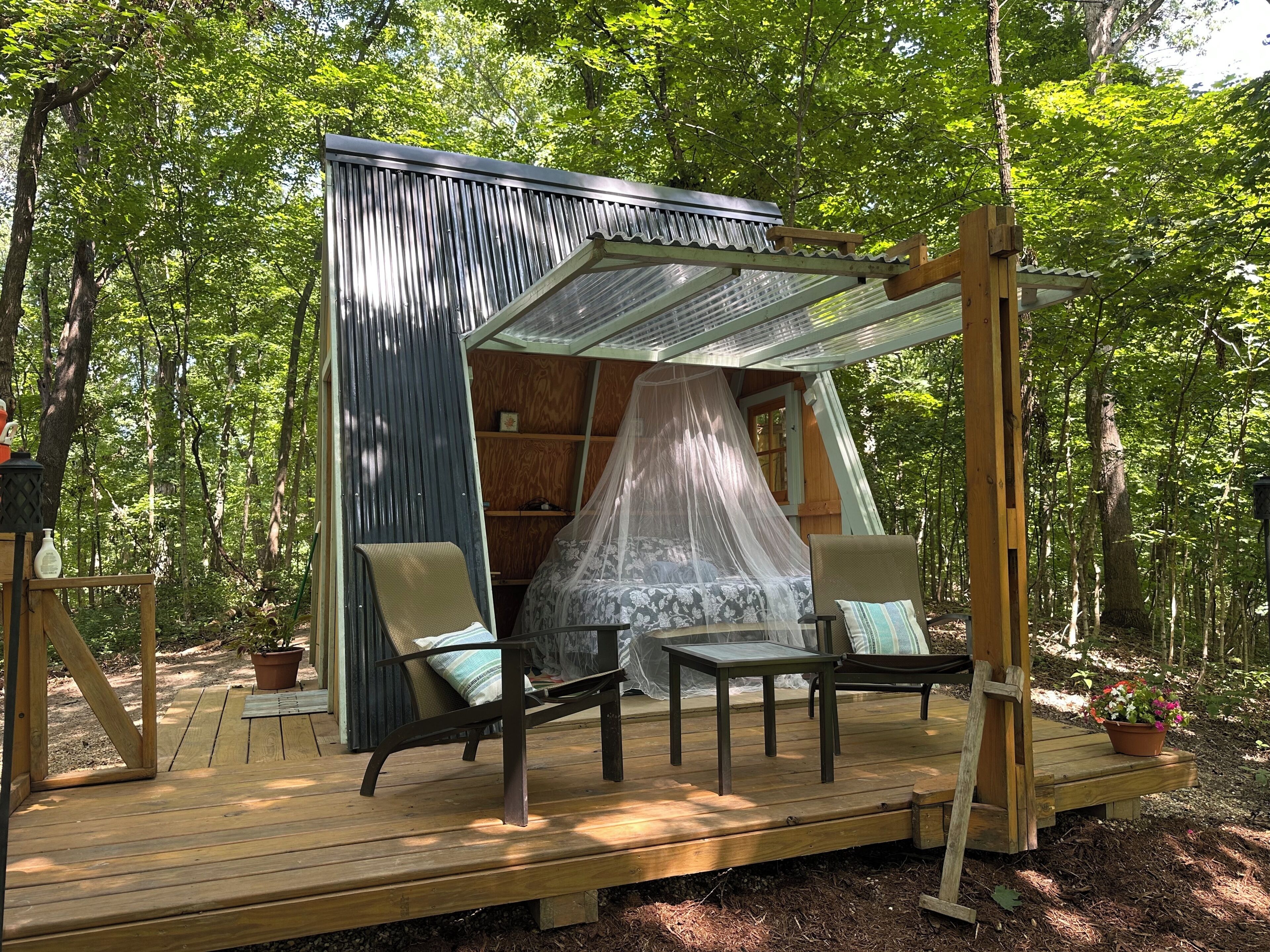 Une terrasse en bois dotée d’un espace salon couvert, d’un petit espace de couchage fermé avec un auvent, et bordée par une forêt.
