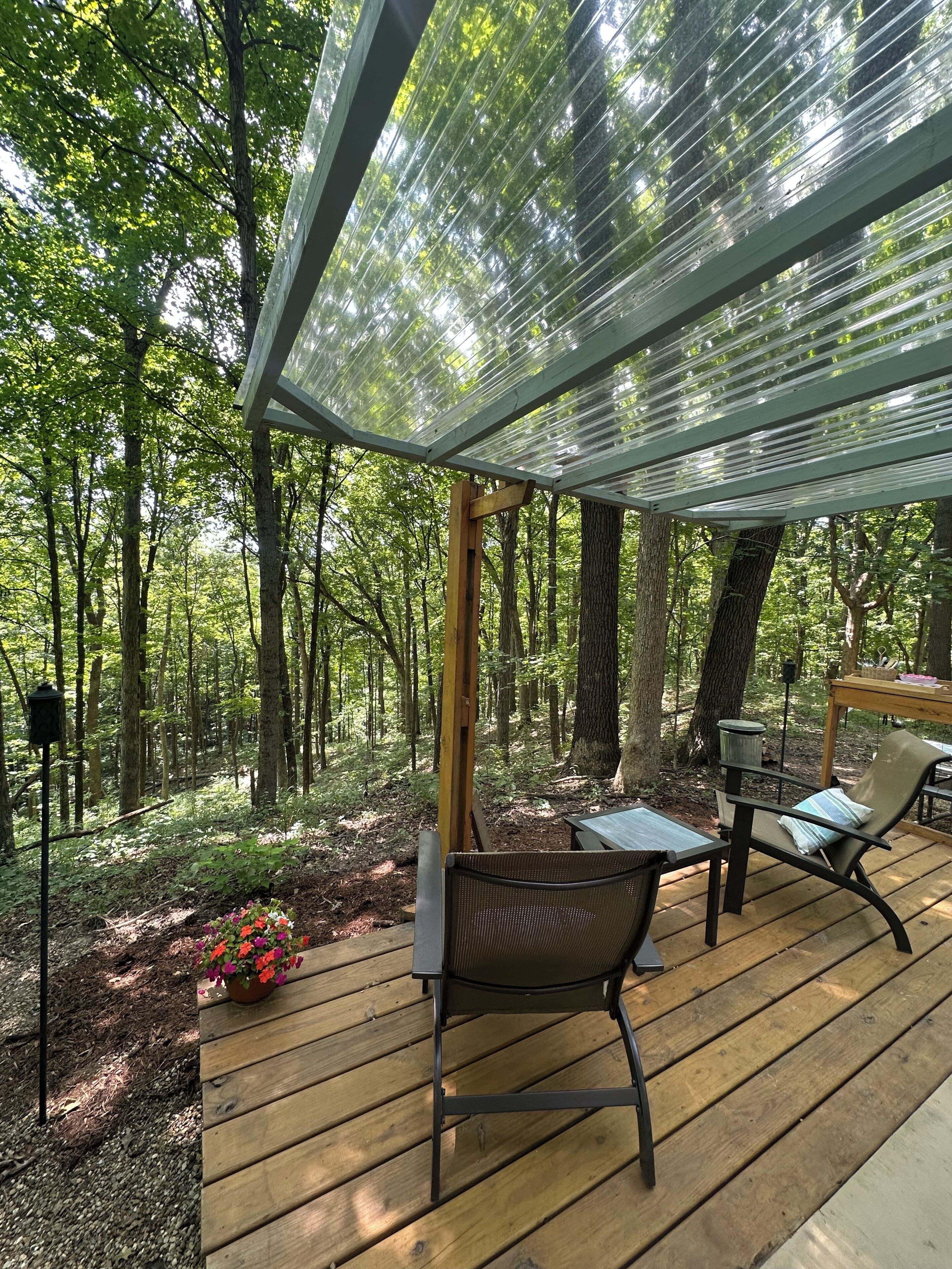 A wooden deck with a glass ceiling, surrounded by a forest. There are two chairs and a table.