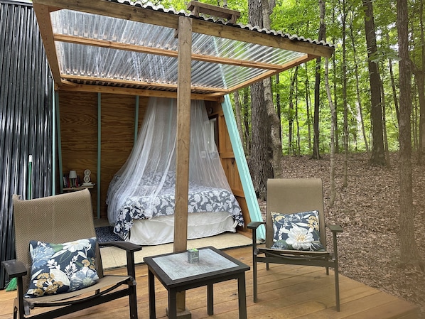 A wooden outdoor seating area with a canopy, a bed with a mosquito net, and a forest backdrop. - A frame getaway atop a ridge in Charleston, Illinois. (Charleston)