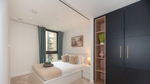 A bedroom with a bed, a window with curtains, a bedside table, a nightstand, a dark wardrobe, and a wall-mounted shelf with decorative items. - Farringdon Lux 2-bed flat w. Balcony - Clerkenwell (London)