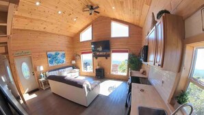 A cozy cabin interior with a wooden ceiling, a sofa, a TV, a kitchen area, and a large window offering a view. - Million Dollar View Home by New River Gorge Park (Cool Ridge)