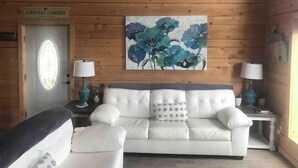 A cozy living room with a white sofa, two side tables with lamps, a floral painting on the wall, and a wooden door with a sign. - Million Dollar View Home by New River Gorge Park (Cool Ridge)