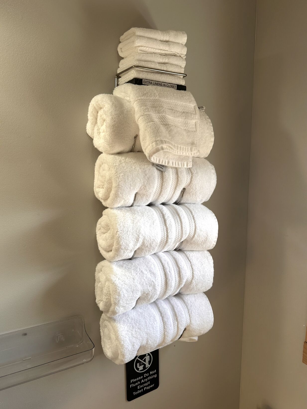 Ein Stapel weiße Handtücher auf einem an der Wand befestigten Ablagefach. Darunter ein Schild mit der Aufschrift 'Bitte nichts außer Toilettenpapier herunterspülen'.