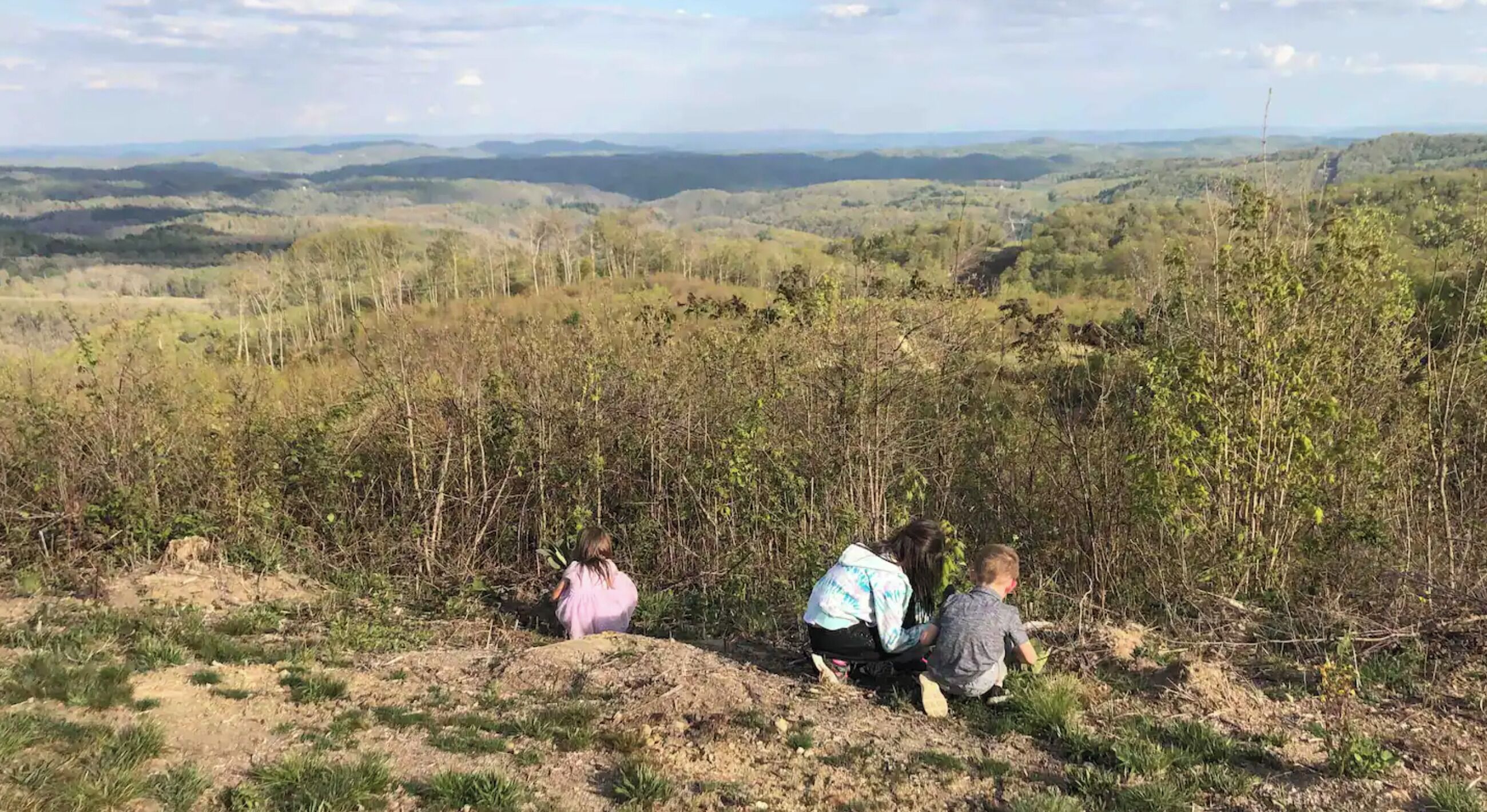 Eine Gruppe von Menschen sitzt auf einem grasbewachsen Hügel mit Blick auf eine bewaldete Landschaft und entfernte Hügel.