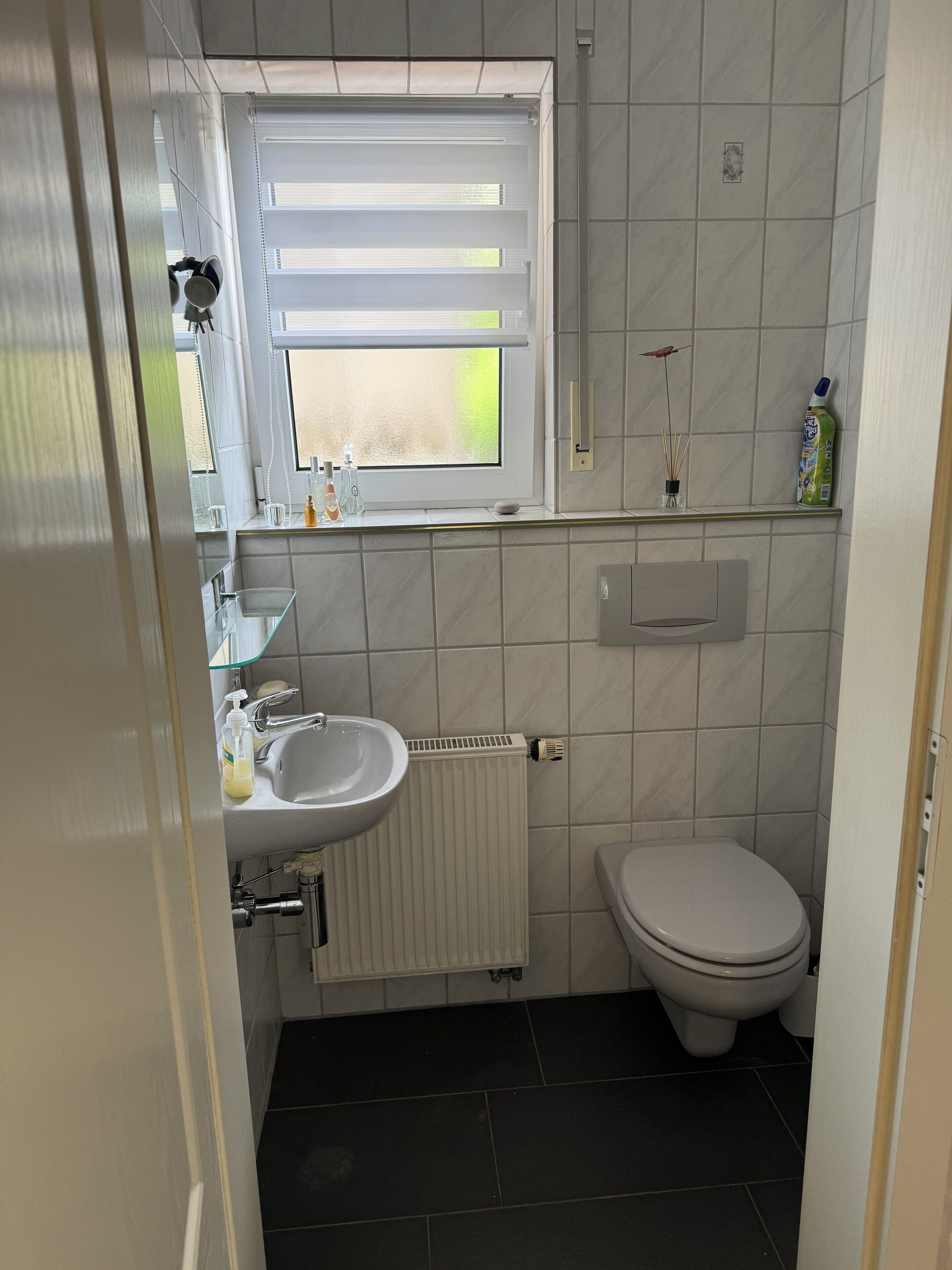 Ein kleines Badezimmer mit einem weißen Waschbecken, einer Toilette und einem Fenster mit Jalousien.