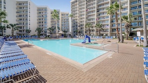 Zona de piscina de hotel con varios niveles de balcones, palmeras y sillas reclinables azules.
