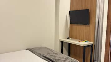 A hotel room with a bed, a desk with a flat-screen TV, a wooden wardrobe, and an air conditioning unit.