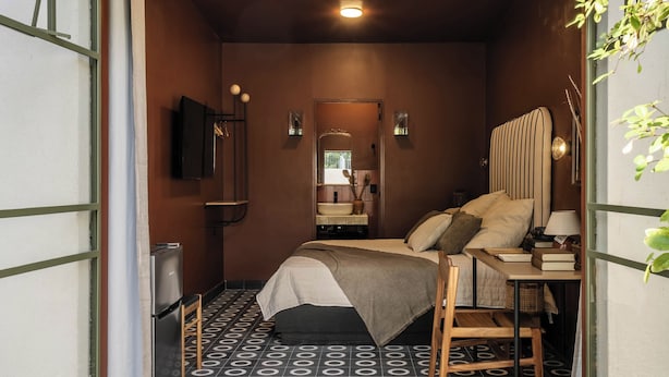 A compact, well-lit bedroom with a bed, bedside table, and a mirror. The room features a patterned floor and a small window.
