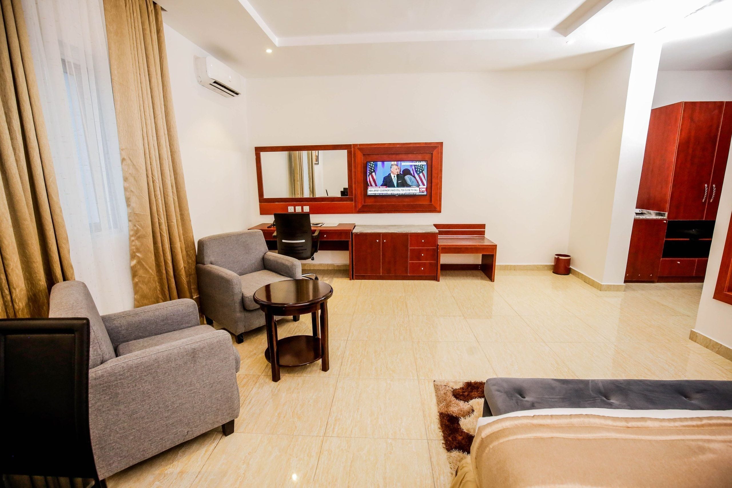 A hotel room with a grey sofa, a small round table, a TV mounted on the wall, and a wooden desk with drawers.