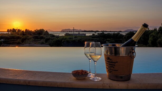 A sunset view from a poolside area with a champagne bottle, two glasses, and a bowl of nuts.