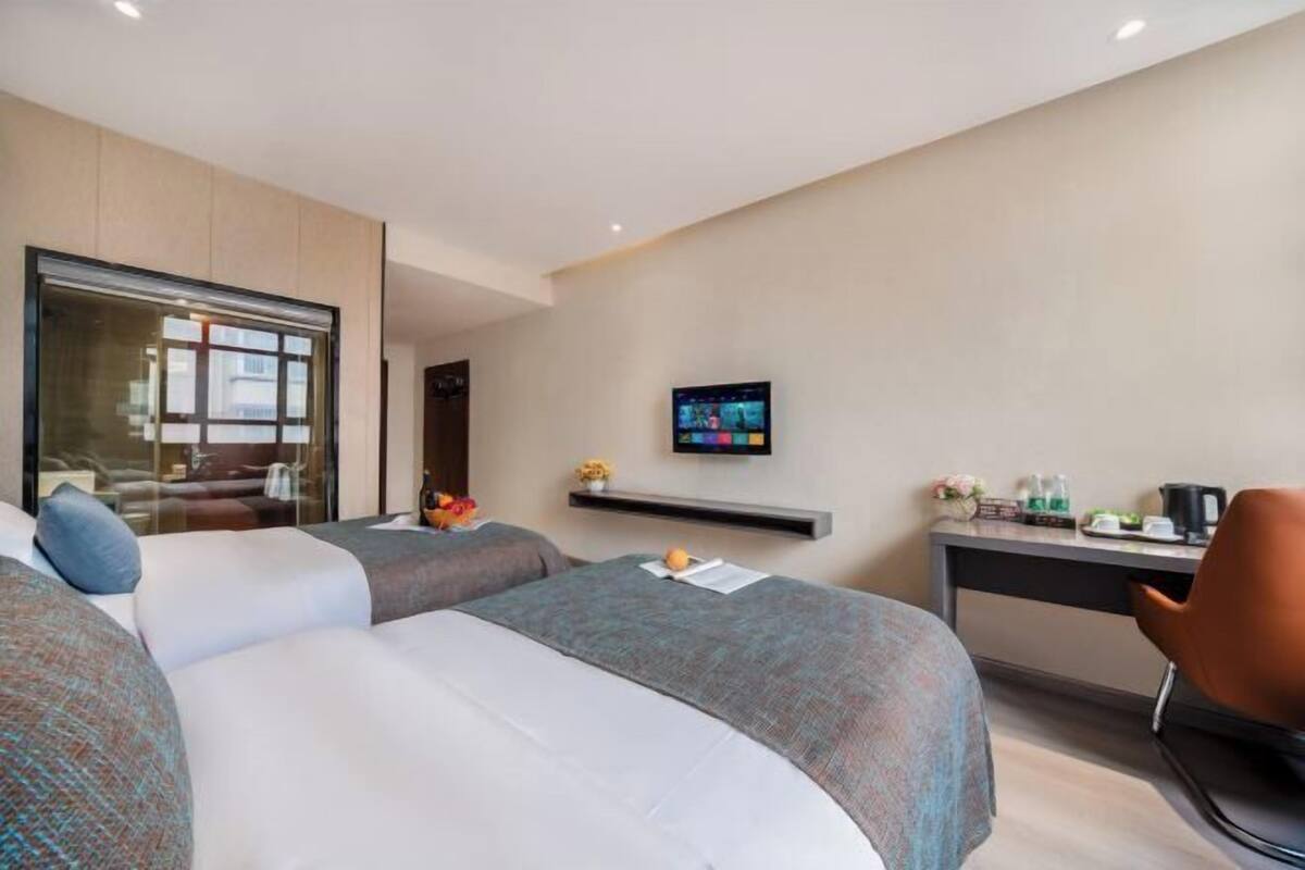A hotel room with two beds, a TV mounted on the wall, a desk with a chair, and a window with a view.