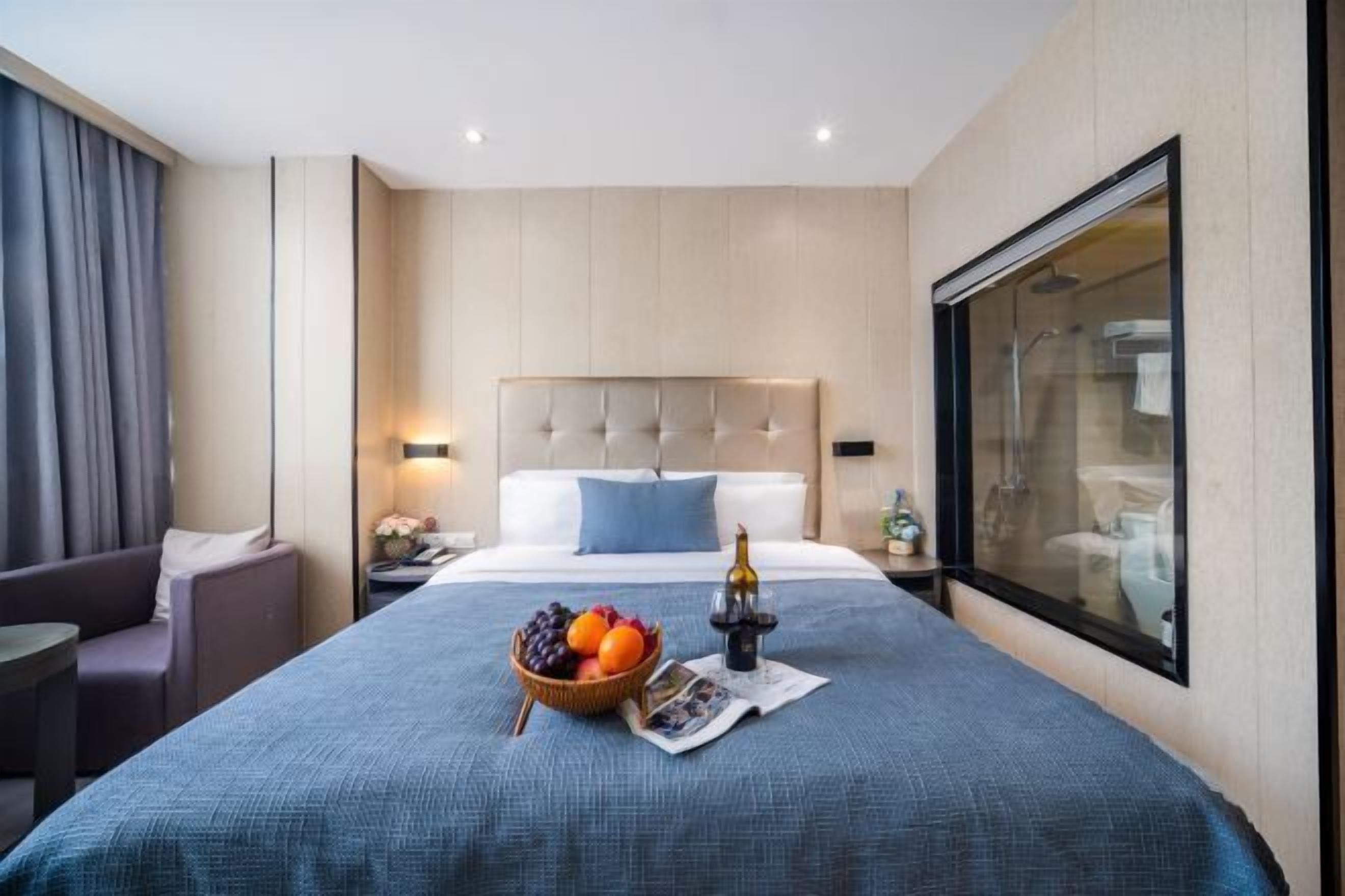 A hotel room with a bed, a bedside table with a lamp, a fruit basket, and a window.