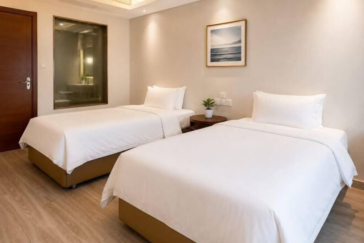 A hotel room with two beds, a wooden floor, a door, a nightstand with a lamp, and a framed picture on the wall.