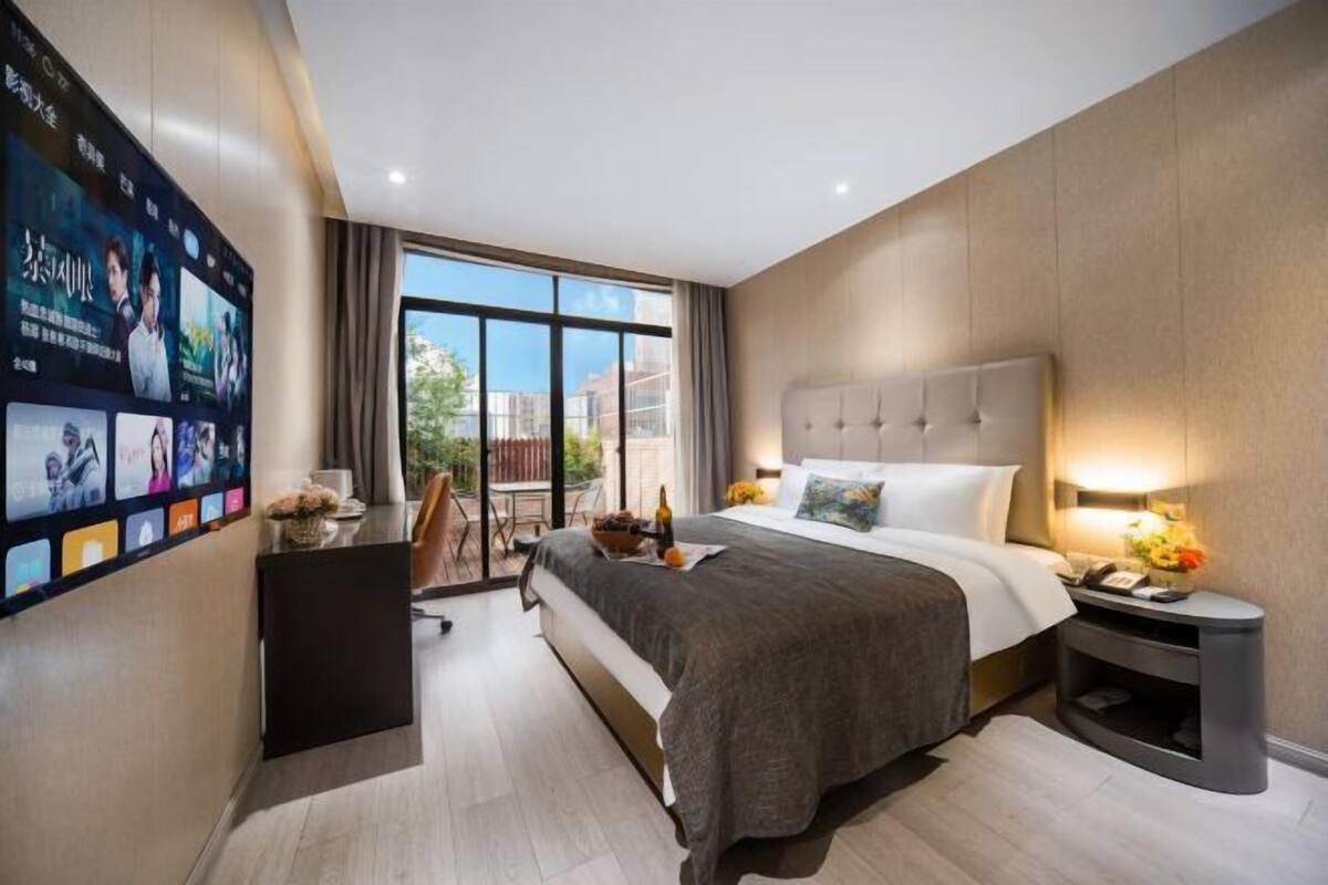 A modern hotel room with a large bed, a desk, a TV mounted on the wall, and a balcony with a view of the city.