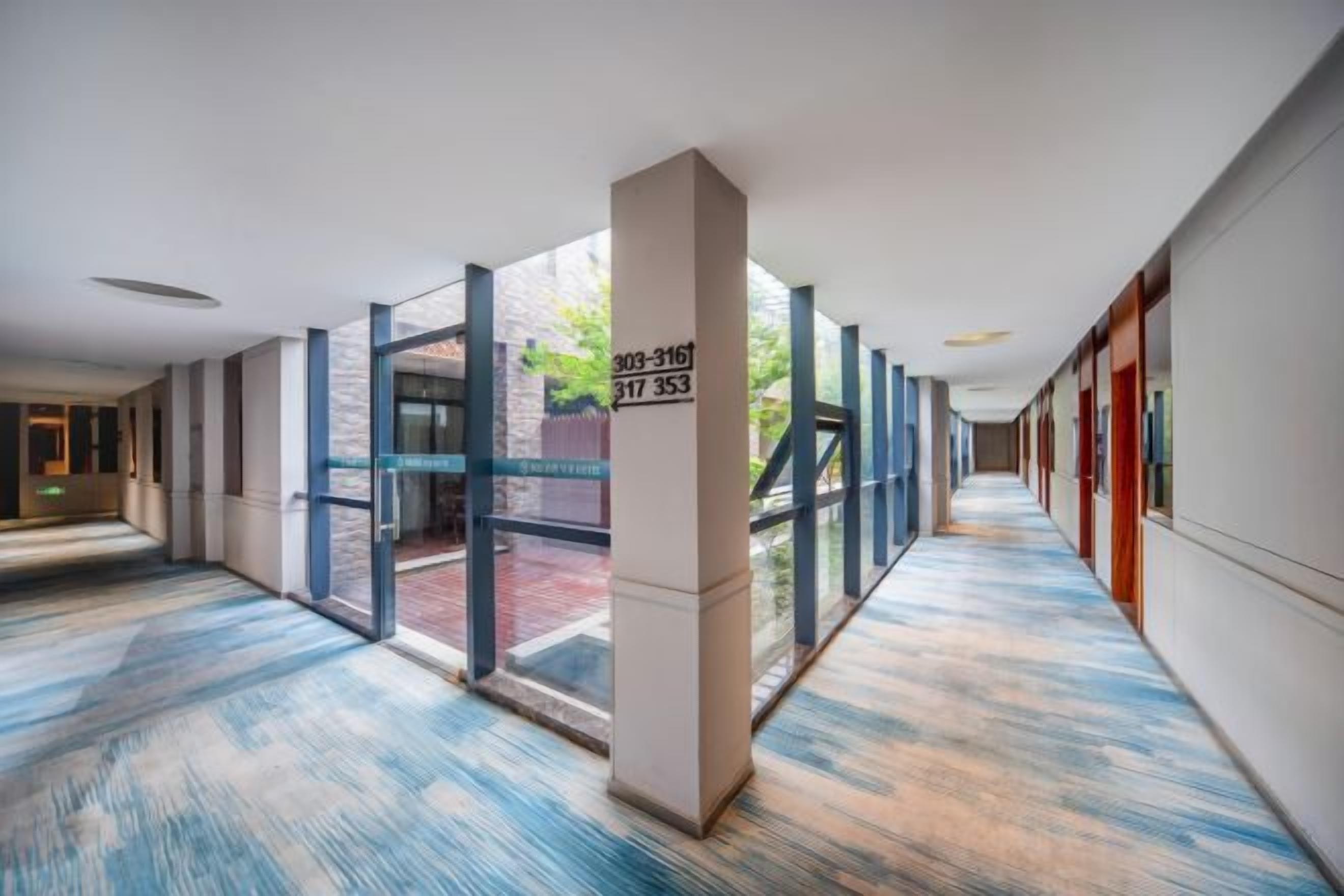 A long corridor with glass doors leading to an outdoor area, a patterned carpet, and a column with a number.