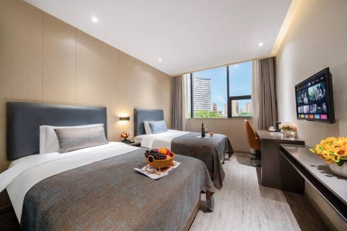 A hotel room with two beds, a desk, a TV, and a window with a city view.