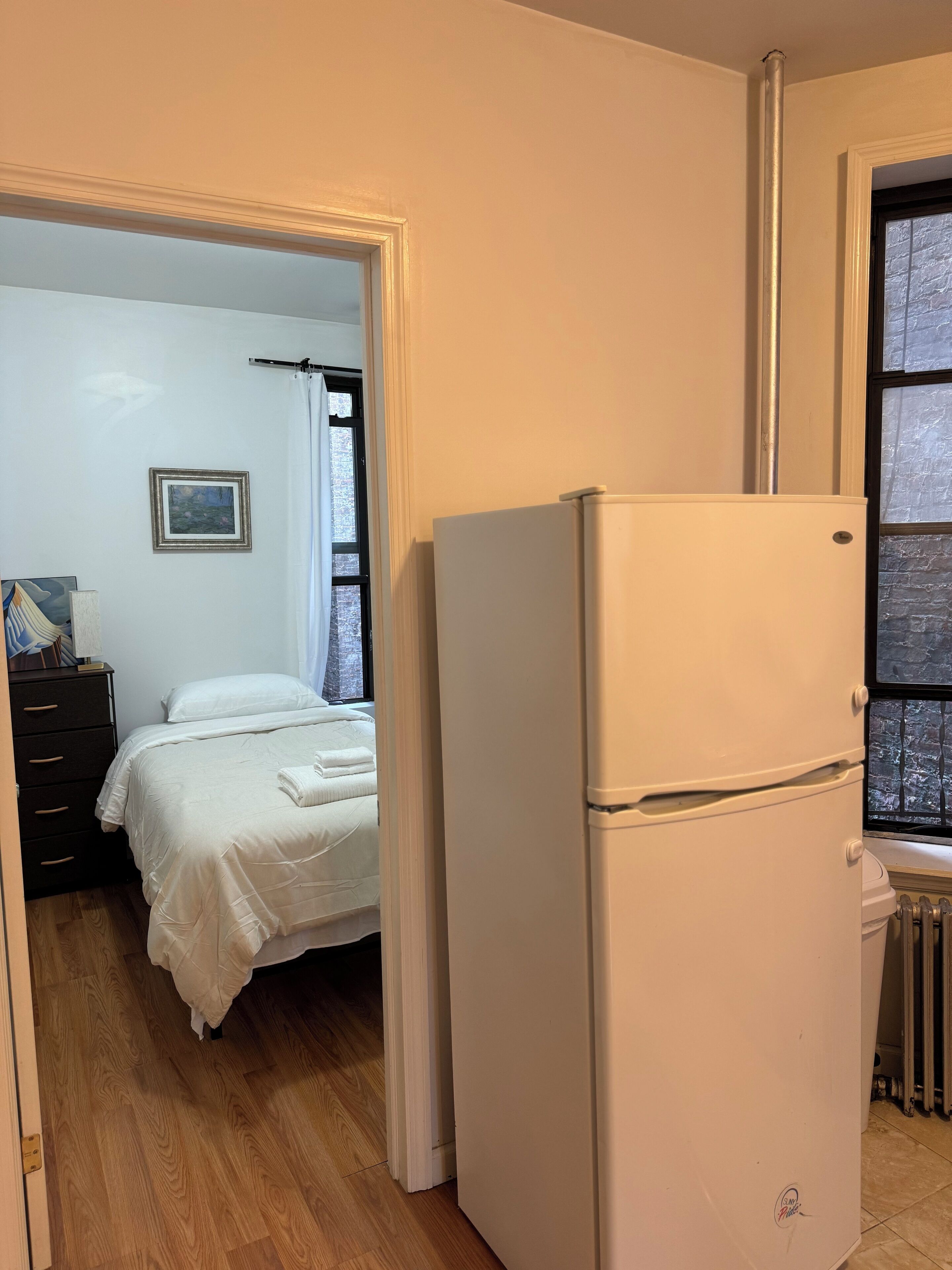 A bedroom with a bed, a white refrigerator, a radiator, and a window with a view of a brick wall.