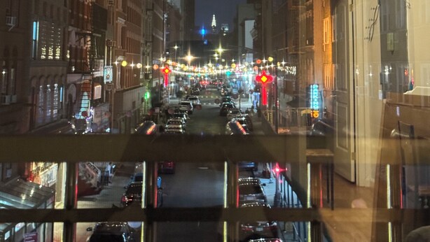 Uitzicht op een verlichte stadstraat 's nachts vanuit een raam, met gebouwen, lantaarns en geparkeerde auto's.