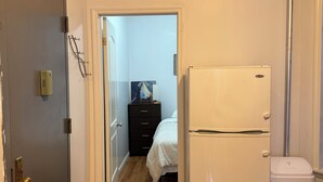 Een smalle gang met een houten vloer die leidt naar een slaapkamer met een bed en een dressoir. Een witte koelkast is in de kamer zichtbaar.