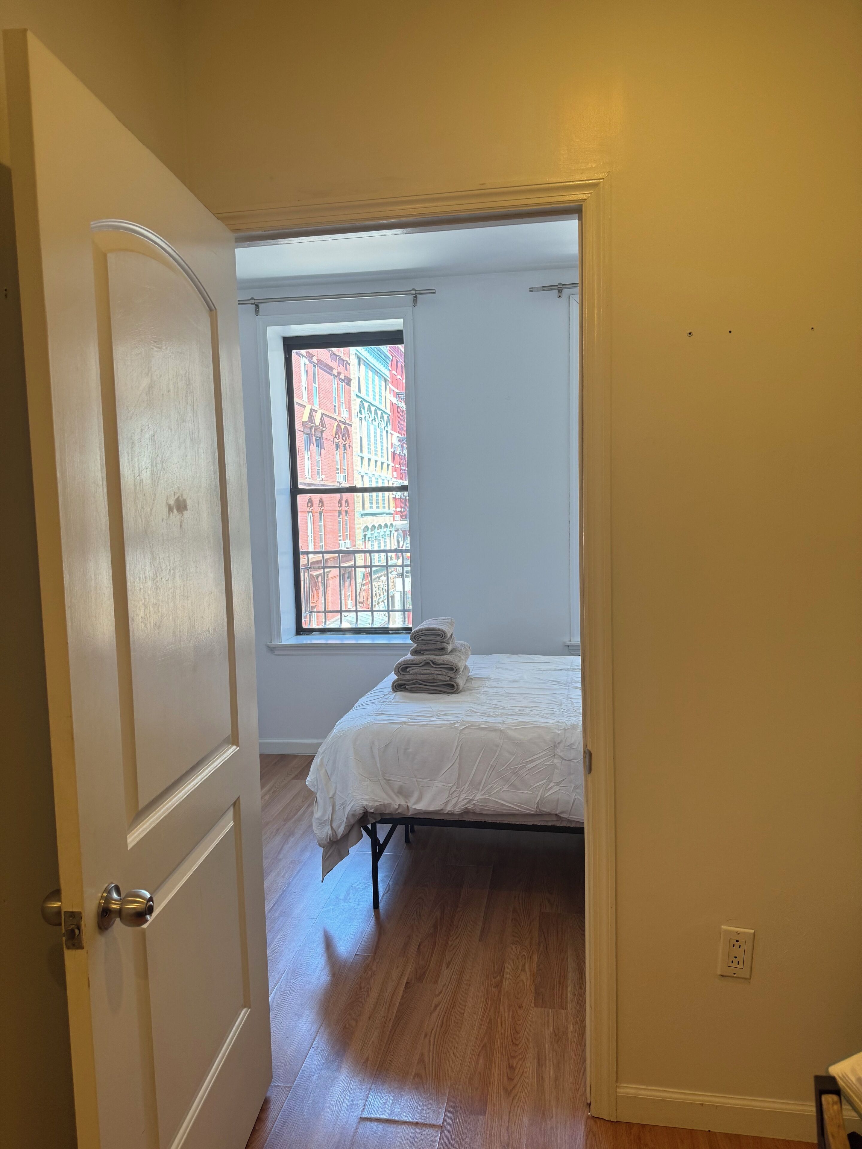 Quarto com cama, janela e vista para edifícios, e uma porta.