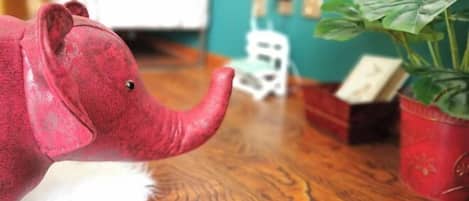 Un elefante de juguete sobre un piso de madera, con una planta en maceta y un objeto blanco pequeño al fondo.