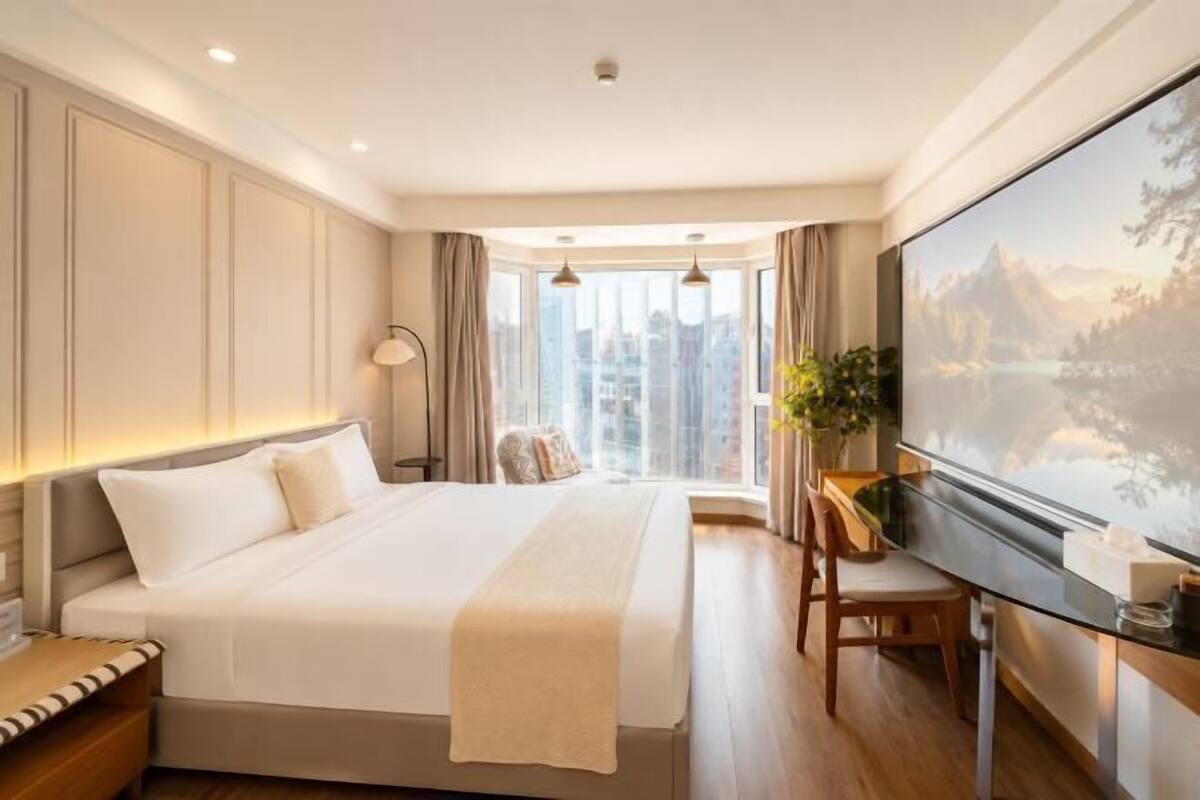 Une chambre d’hôtel avec un grand lit, un bureau avec une chaise, une grande fenêtre donnant sur une montagne et sur la ville, et un tableau accroché au mur.