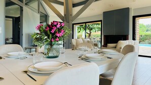 A modern dining area with a long table set for a meal, surrounded by white chairs, and a vase of pink flowers.