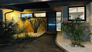 A modern house entrance with a wooden fence, a pathway leading to a dark door, and a garden area with plants and pebbles.