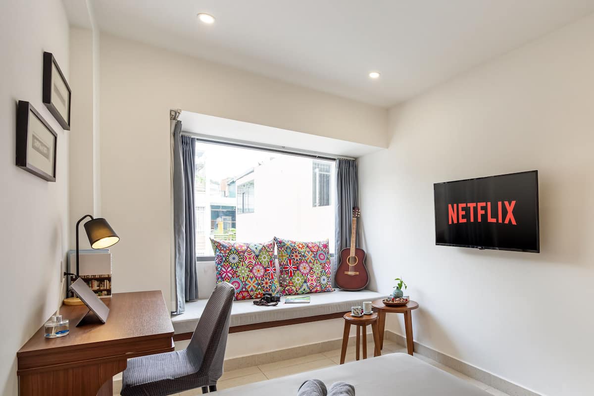 Moderni hotellihuone, jossa on suuri ikkuna, työpöytä lampun kanssa, tuoli, sänky, pieni pöytä, jossa on kasvi, ja televisio, jossa näkyy teksti ”NETFLIX”.
