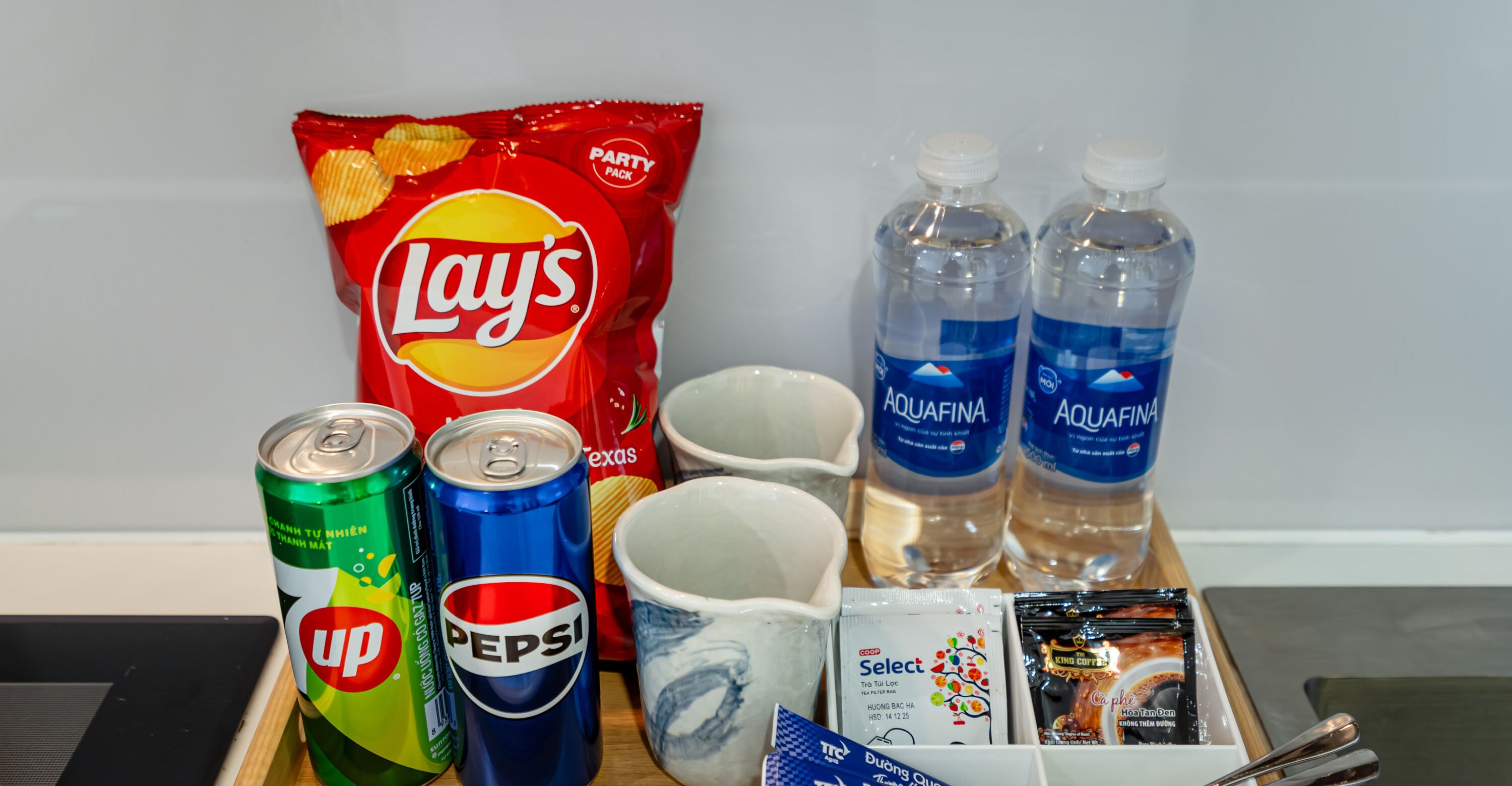 Una bandeja con papas fritas Lay's, latas de Pepsi, botellas de agua Aquafina, una taza blanca, una pequeña bolsita de crema para café y una bolsita de café instantáneo.