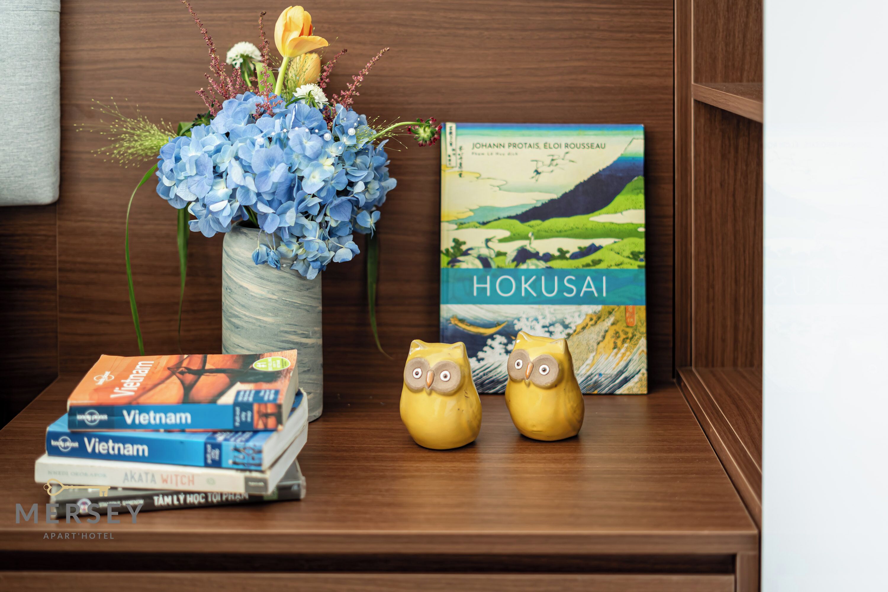 A wooden shelf with a vase of blue flowers, a book titled 'Hokusai', and two owl figurines.