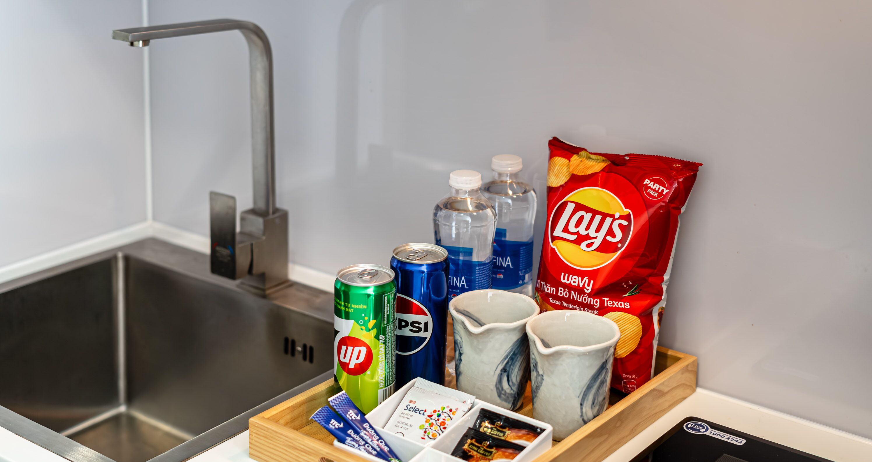 Eine Küchenarbeitsplatte mit Spüle, einer Dose 'Up'-Energydrink, einer Dose 'Pepsi', zwei Wasserflaschen, einer Tüte 'Lay's'-Kartoffelchips und zwei Keramiktassen.