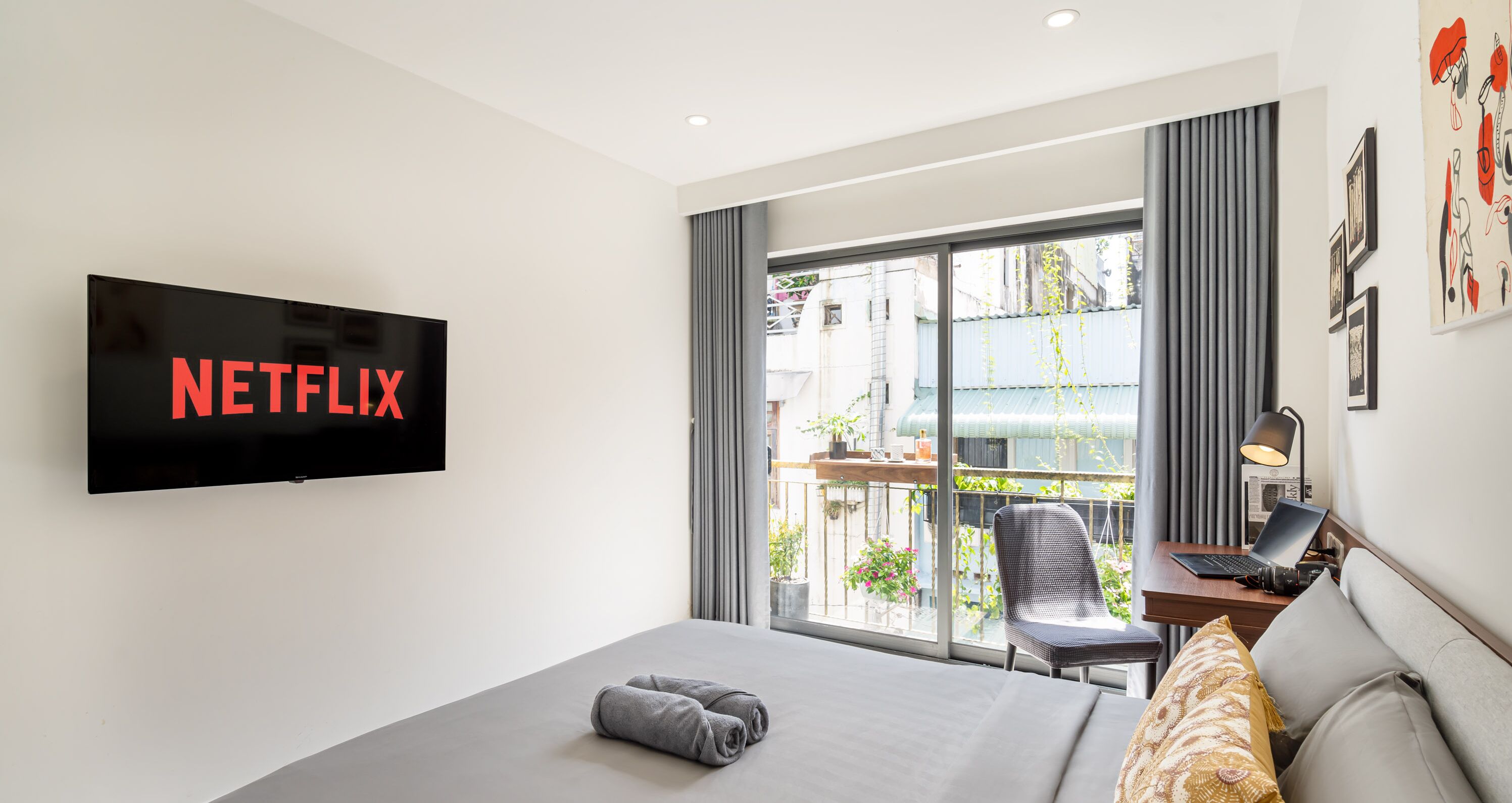 A modern hotel room with a large bed, a desk with a laptop, a television displaying "NETFLIX," and a view of the outside balcony.